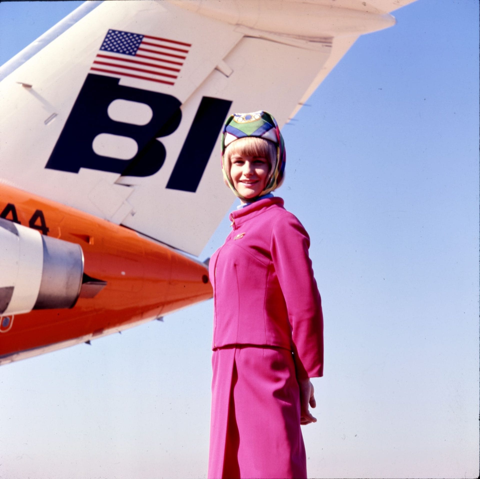 Une hôtesse de l'air en uniforme rose devant un avion orange Braniff International